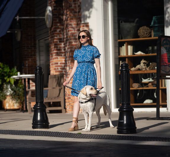 A young girl wearing a blue dress and sunglasses walking with a yellow Labrador guide dog in front of a brick building with large windows.